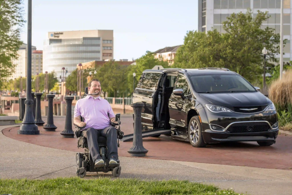 A man in a power wheelchair is enjoying a sunny day near a waterfront promenade. Behind him, a black wheelchair-accessible van with an extended ramp is parked. The background features modern office buildings and trees, suggesting an urban environment.