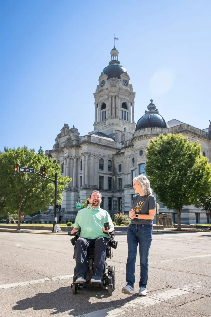A man in a power wheelchair, wearing a light green polo shirt, laughs while conversing with a woman in a casual t-shirt and jeans. They are standing in front of a historic courthouse with a domed roof and intricate architecture. The blue sky and greenery add vibrancy to the scene.