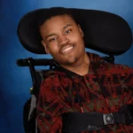 A close-up studio portrait of the young man in a wheelchair, wearing a patterned sweater. He is smiling warmly against a professional blue backdrop.