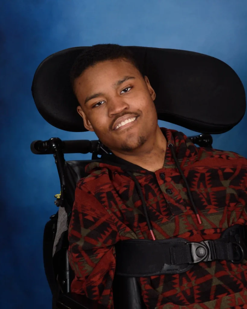 A close-up studio portrait of the young man in a wheelchair, wearing a patterned sweater. He is smiling warmly against a professional blue backdrop.