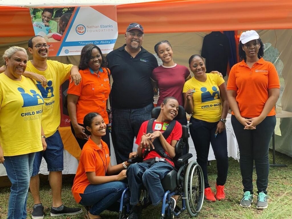 A diverse group of people, wearing yellow and orange event shirts, gathers around the young man in a wheelchair at an outdoor advocacy event. A banner for the Nathan Ebanks Foundation, featuring its logo and slogan, is displayed in the background.