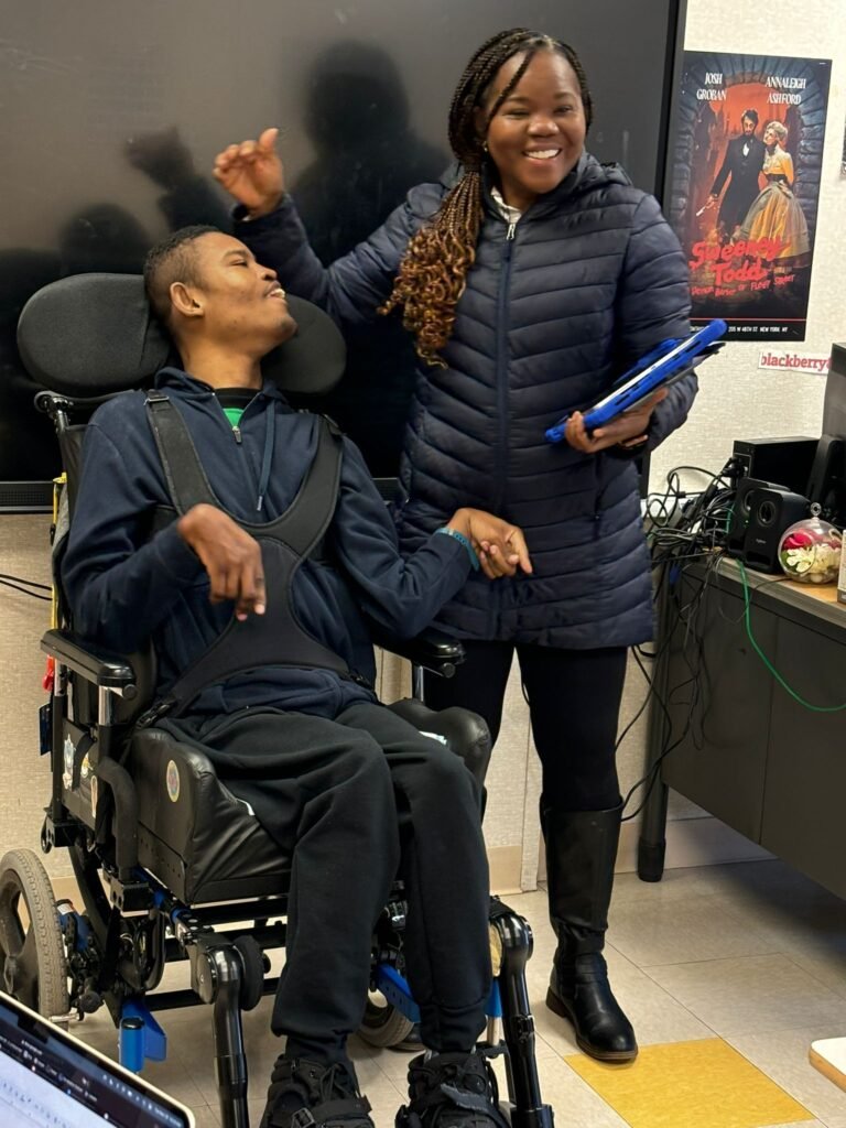 The young man in a wheelchair is interacting with a woman standing next to him. She is holding a tablet, smiling, and appears to be facilitating a discussion or presentation. A large screen and various equipment are visible in the background.