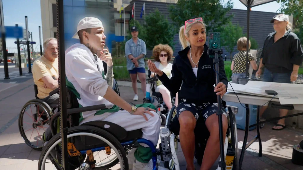 A scene from PUSH showing a group of individuals, some in wheelchairs, engaged in a discussion outdoors. A woman with short platinum blonde hair, wearing a black outfit and holding a smartphone on a tripod, appears to be leading the conversation, while a man in a white cap and hoodie listens attentively. Other people are visible in the background, including a child and a woman in casual attire.