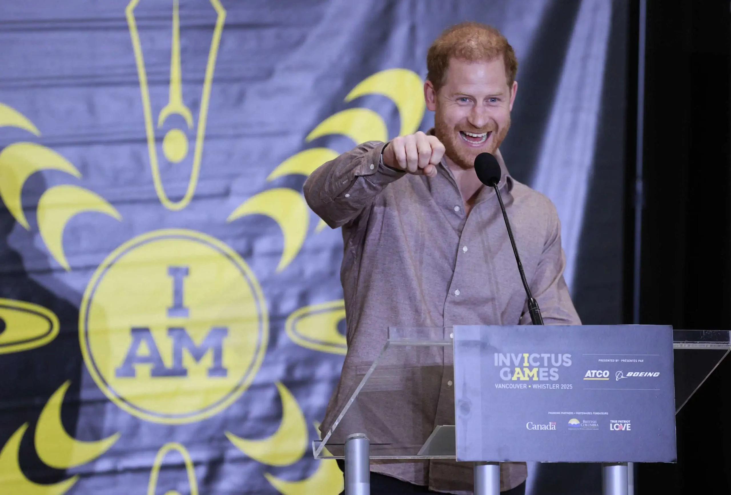 Prince Harry, wearing a light brown button-up shirt, speaks at a podium during an Invictus Games event. He has a broad smile and is gesturing with his fist towards the audience. Behind him, a banner displays the Invictus Games logo with the words "I AM" in bold yellow letters. The podium sign indicates the event is promoting the Invictus Games Vancouver Whistler 2025, with sponsorship logos including ATCO, Boeing, and the Canadian government. Photo Credit to Invictus Games Vancouver Whistler 2025