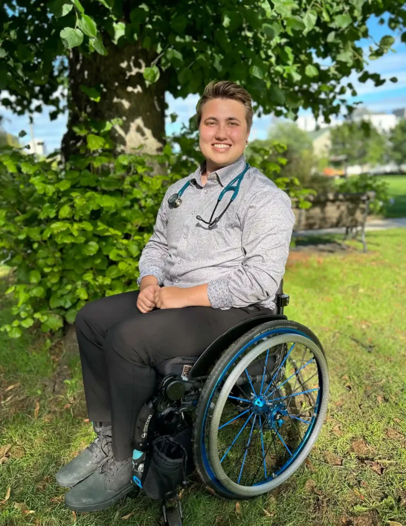 A young man with short brown hair and light skin is sitting in a wheelchair with blue accents in a grassy park. He is wearing a light gray button-up shirt and black pants, with a stethoscope around his neck. He is smiling at the camera, and the background features a tree with lush green leaves and a park bench.