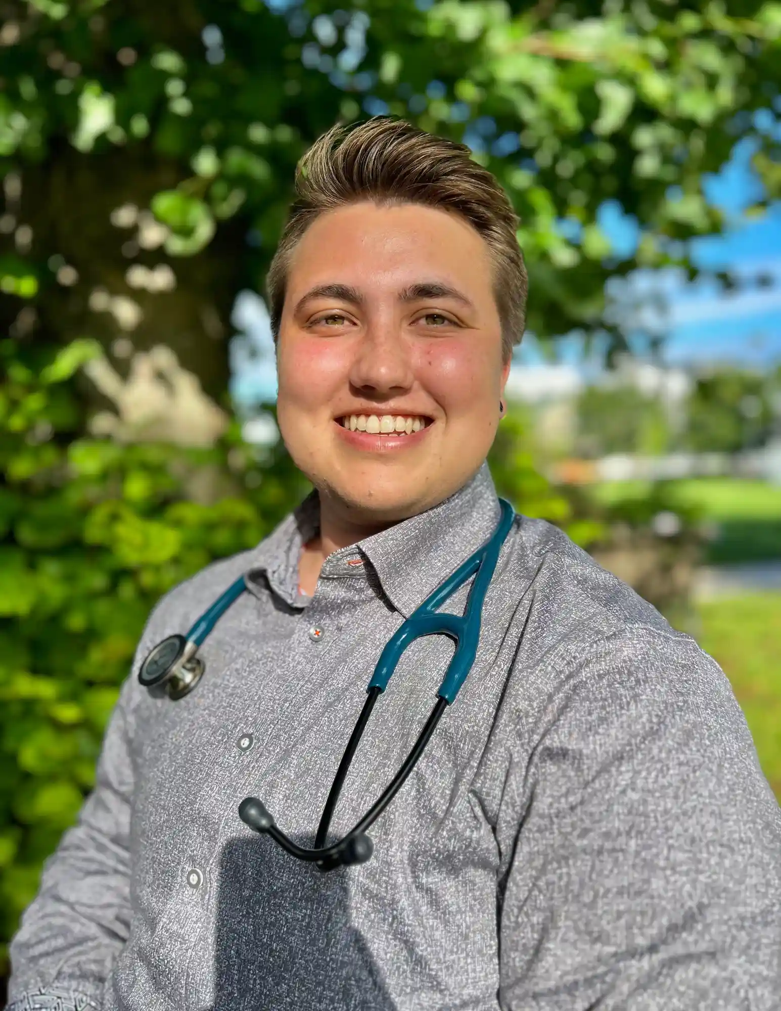 A close-up portrait of a young man with short brown hair and light skin, wearing a light gray button-up shirt and a stethoscope around his neck. He has a broad smile, and the background consists of a tree with green leaves and a bright blue sky.