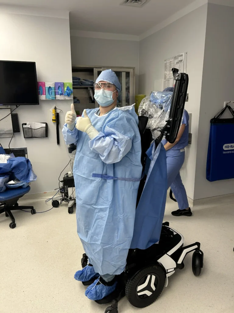 A young man is in a medical operating room, wearing blue surgical scrubs, a cap, a mask, and gloves. He is using a standing power wheelchair, giving two thumbs up. The room contains medical equipment and a storage cabinet in the background.