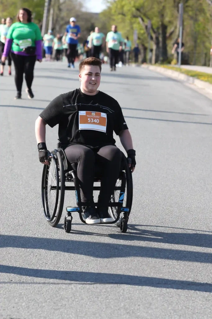 A young man is participating in a race, using a sleek black racing wheelchair. He is wearing a black athletic outfit, and a race bib with the number "5340" is pinned to his shirt. Other runners in the background are jogging along a paved road.