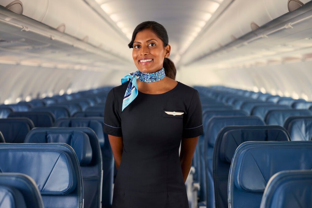 A smiling flight attendant wearing a blue uniform and scarf stands in the aisle of an airplane with rows of empty seats behind her.