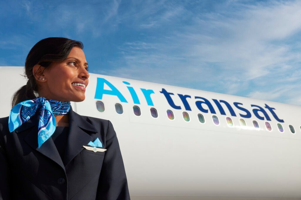 A smiling Air Transat flight attendant is captured in a professional uniform in front of an Air Transat aircraft. She wears a dark navy blazer with a silver wing-shaped pin and a blue pocket square, complemented by a stylish blue patterned scarf tied around her neck. The aircraft in the background prominently displays the "Air Transat" logo on its white fuselage, with a row of oval windows reflecting sunlight. The backdrop features a bright blue sky with wispy clouds.