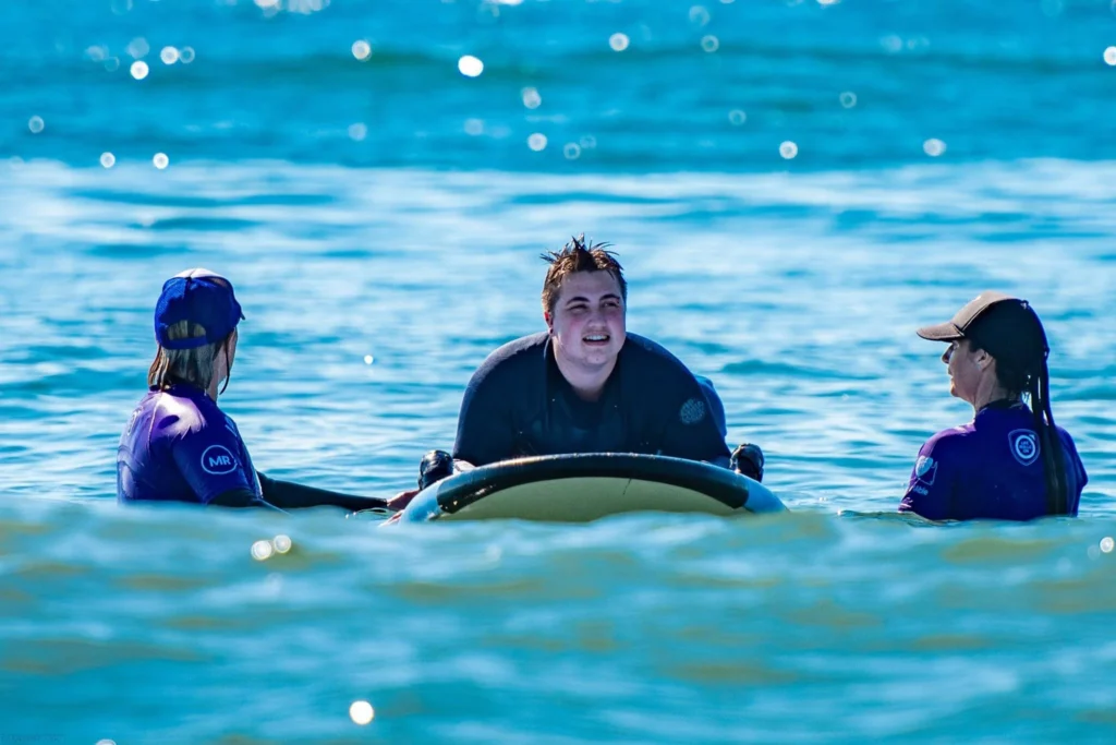 young man is in the ocean, learning to surf with the assistance of two instructors wearing purple shirts. He is lying on a surfboard, smiling, with wet hair. The water is a bright shade of blue, reflecting sunlight. Photo credits to Dave Morash.