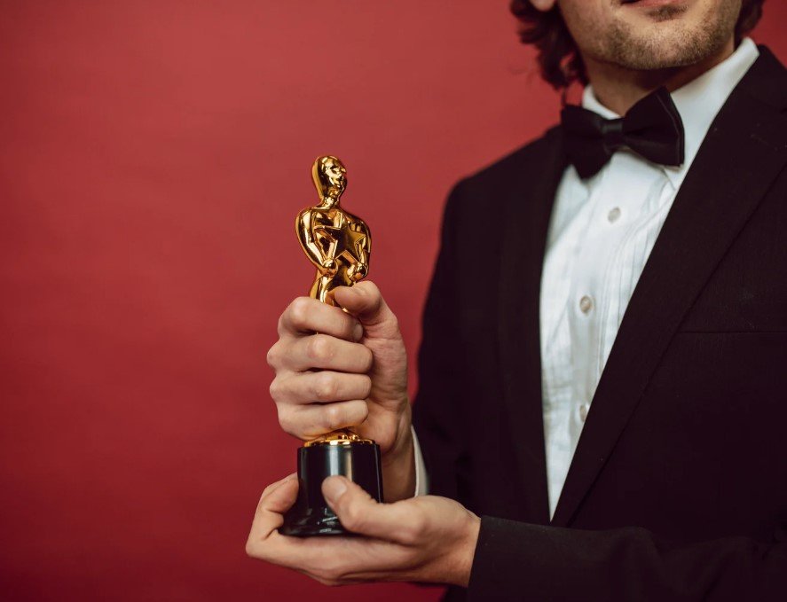 A man dressed in a black tuxedo with a bow tie is holding a golden trophy in both hands. The trophy features a figure holding a star, resembling an award for excellence in film or entertainment. The background is a deep red, adding to the prestigious and celebratory atmosphere.