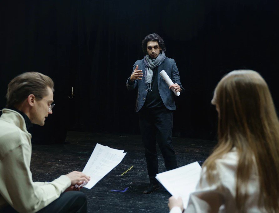 A theatrical director, dressed in a dark blazer and scarf, stands on a dimly lit stage, gesturing with one hand while holding a rolled-up script in the other. Two actors, a man and a woman, are seated in the foreground, attentively reviewing their scripts. The stage has a dark background, emphasizing the focused atmosphere of a rehearsal.