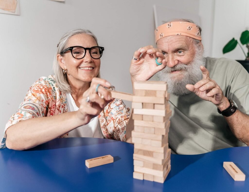 An elderly woman with gray hair, glasses, and a colorful patterned cardigan is smiling while carefully removing a wooden block from a Jenga tower. Beside her, an elderly man with a white beard, wearing a light green shirt, an orange bandana, and a black wristwatch, is playfully observing the game with a focused expression. They are sitting at a blue table, engaged in the game, with wooden blocks scattered around. The background features a light-colored wall with minimal decorations and a green plant.
