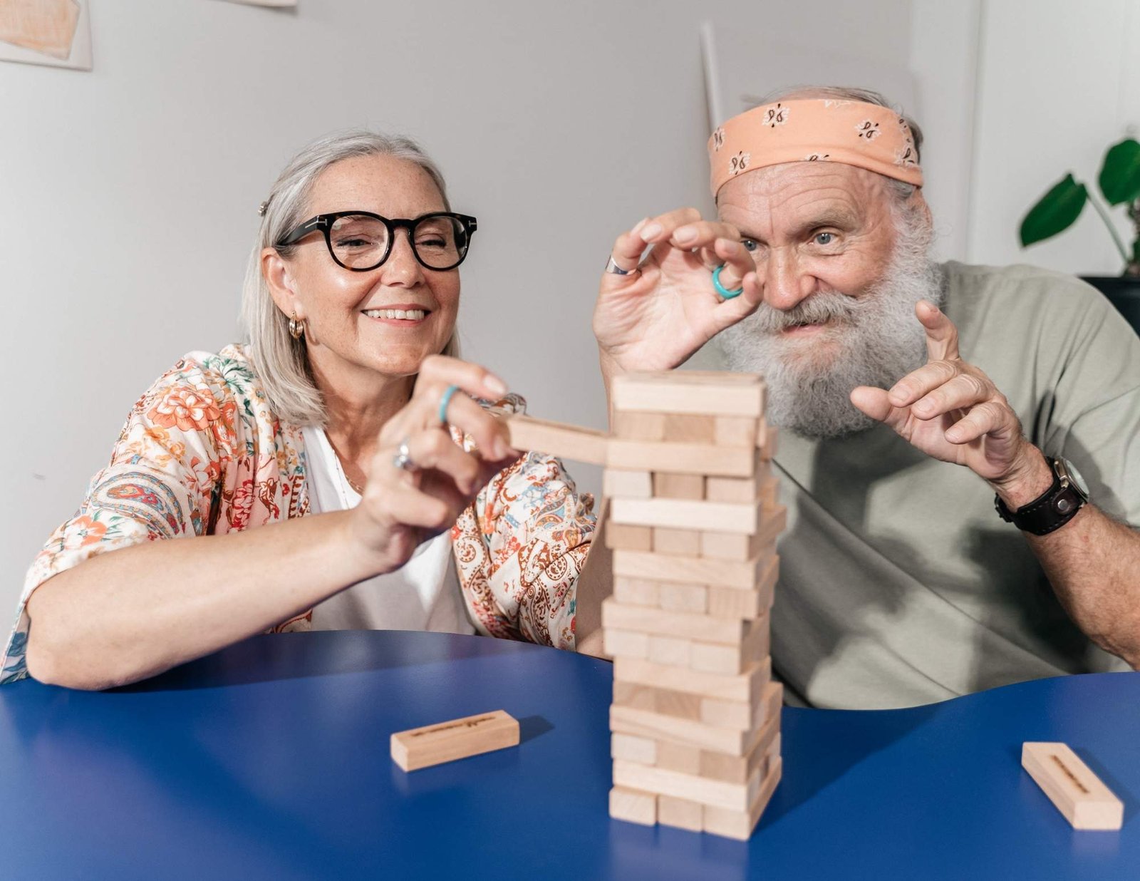 An elderly woman with gray hair, glasses, and a colorful patterned cardigan is smiling while carefully removing a wooden block from a Jenga tower. Beside her, an elderly man with a white beard, wearing a light green shirt, an orange bandana, and a black wristwatch, is playfully observing the game with a focused expression. They are sitting at a blue table, engaged in the game, with wooden blocks scattered around. The background features a light-colored wall with minimal decorations and a green plant.