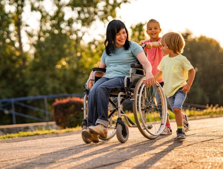A woman in a wheelchair is outdoors on a paved path, smiling and interacting with two young children. One child is holding her hand while running alongside her, and the other is playfully pushing her wheelchair from behind. The setting is warm and sunny, with trees and greenery in the background, creating a joyful and inclusive atmosphere.