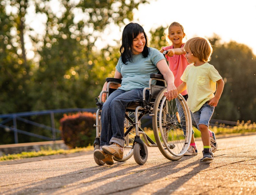 A woman in a wheelchair is outdoors on a paved path, smiling and interacting with two young children. One child is holding her hand while running alongside her, and the other is playfully pushing her wheelchair from behind. The setting is warm and sunny, with trees and greenery in the background, creating a joyful and inclusive atmosphere.