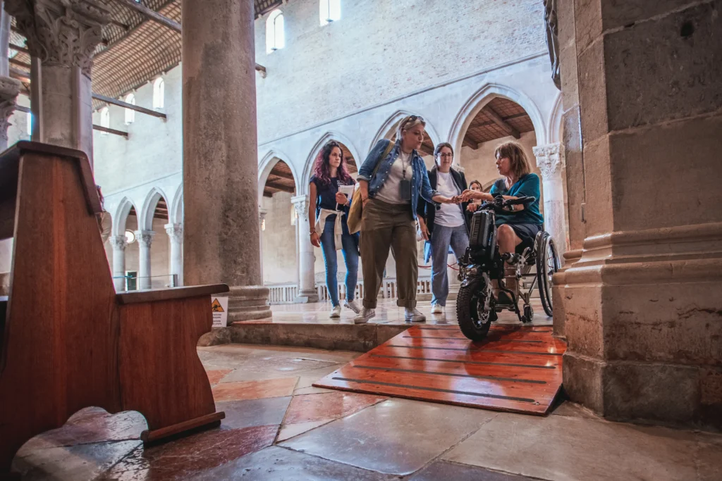 A group of people assists a woman in a wheelchair navigating a ramp inside an architectural space with arches and wooden accents.