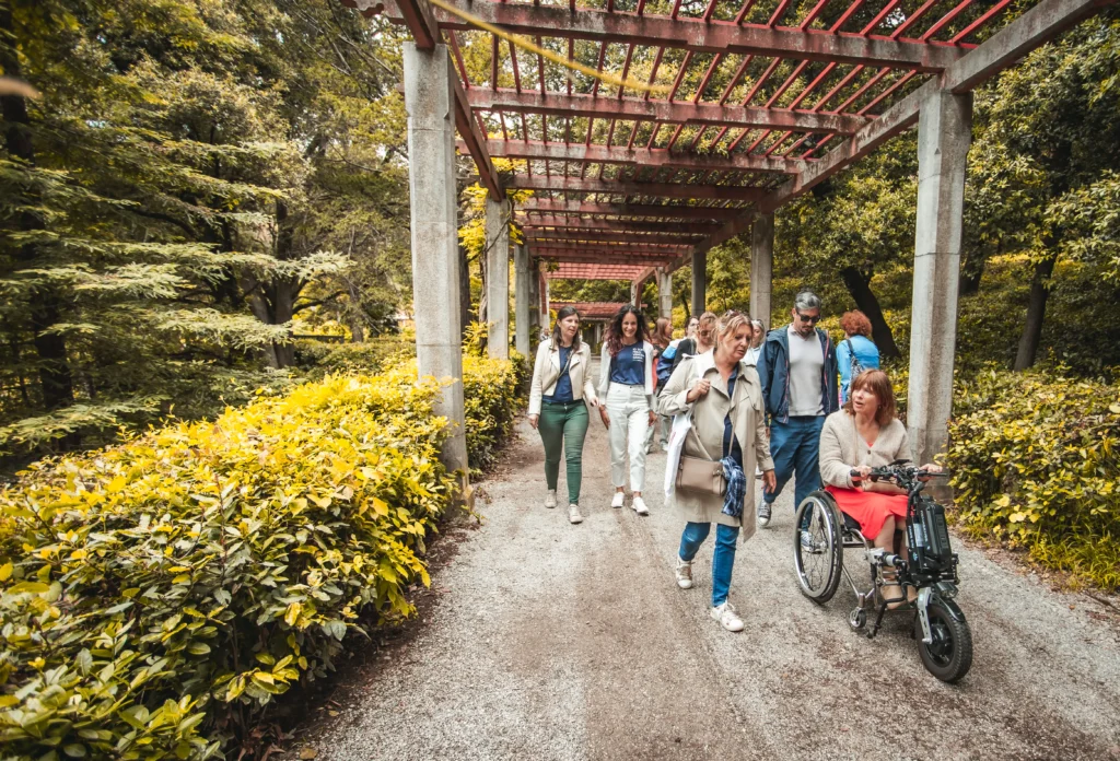 A diverse group of people walking on a path surrounded by lush greenery and a pergola, enjoying a day outdoors.