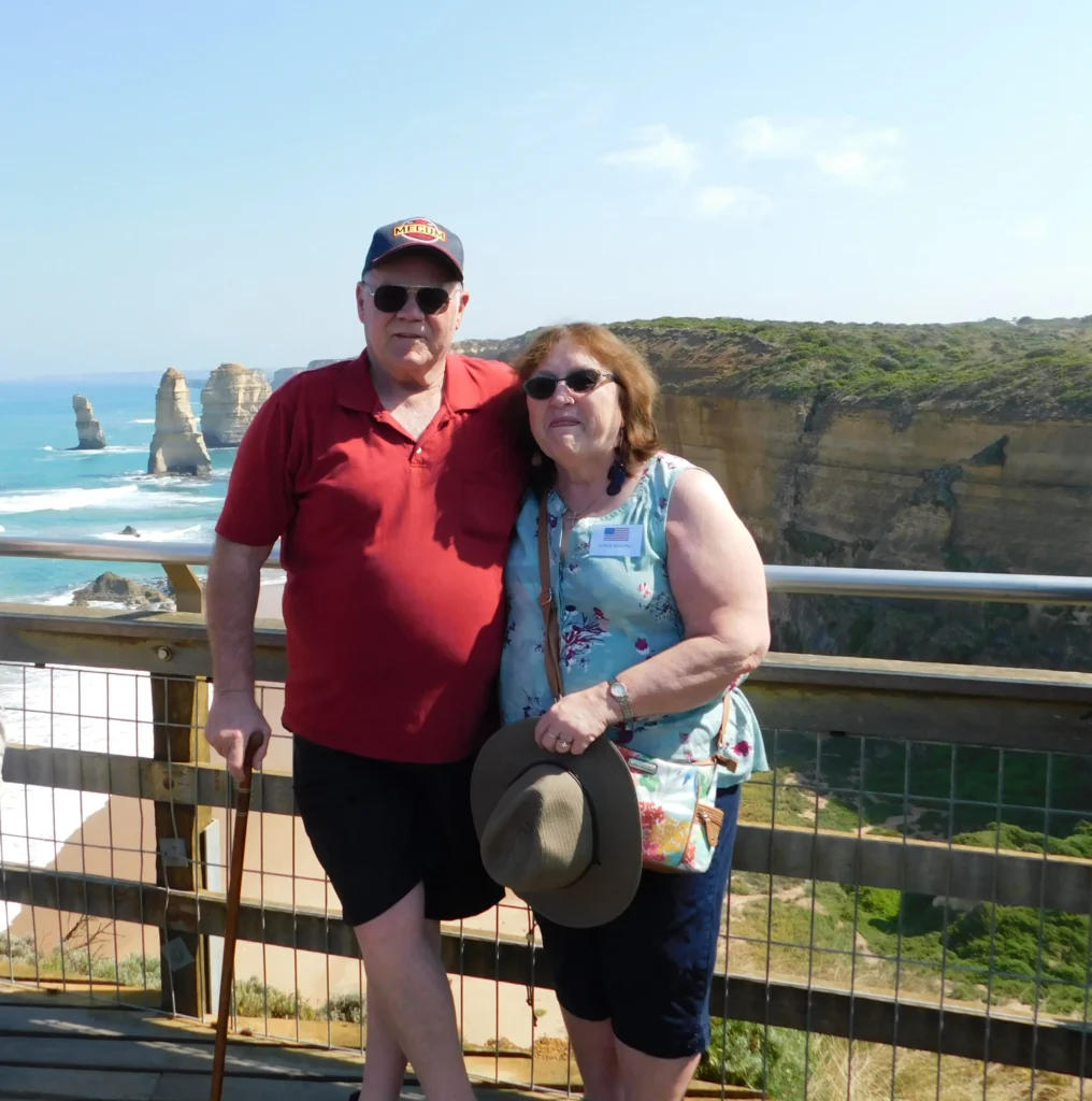 A couple stands together by a scenic viewpoint overlooking the ocean and rugged cliffs, with unique rock formations in the background.