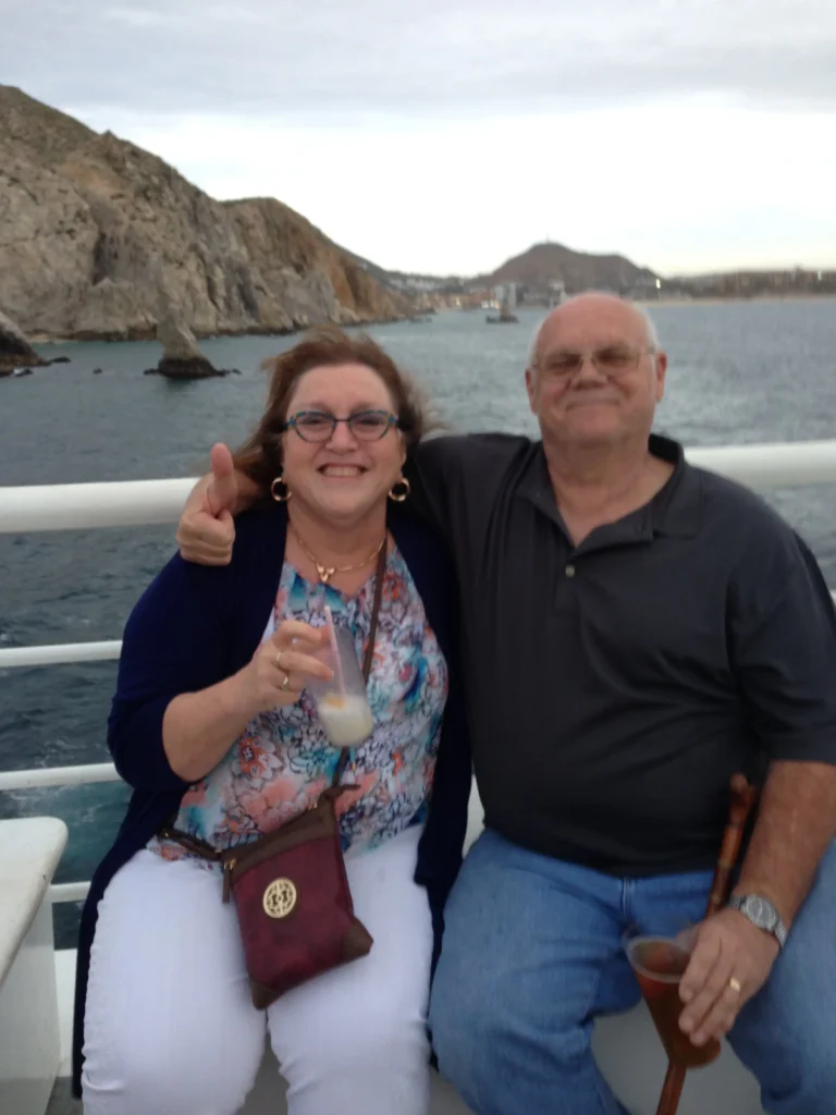 A couple enjoys drinks on a boat, with rocky cliffs and a calm sea in the background during a cloudy evening.