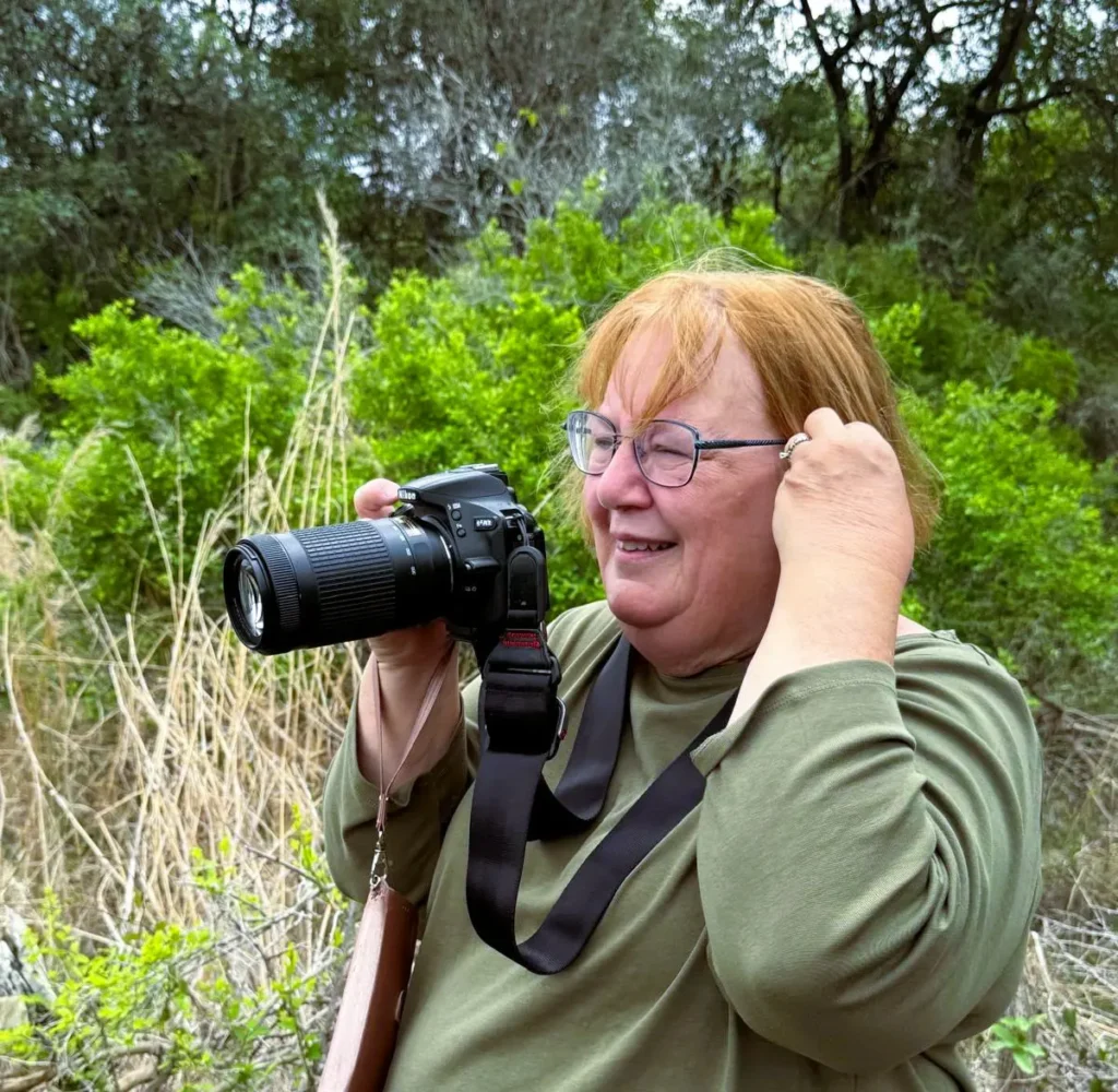 A middle-aged woman with light skin, reddish-brown hair, and glasses is standing outdoors in a green, natural environment. She is wearing an olive-green long-sleeve shirt and adjusting her hair while holding a DSLR camera with a long zoom lens. The camera strap hangs around her neck, and she is smiling. The background consists of green foliage, dry grass, and trees. The setting appears to be a nature reserve or a forested area.