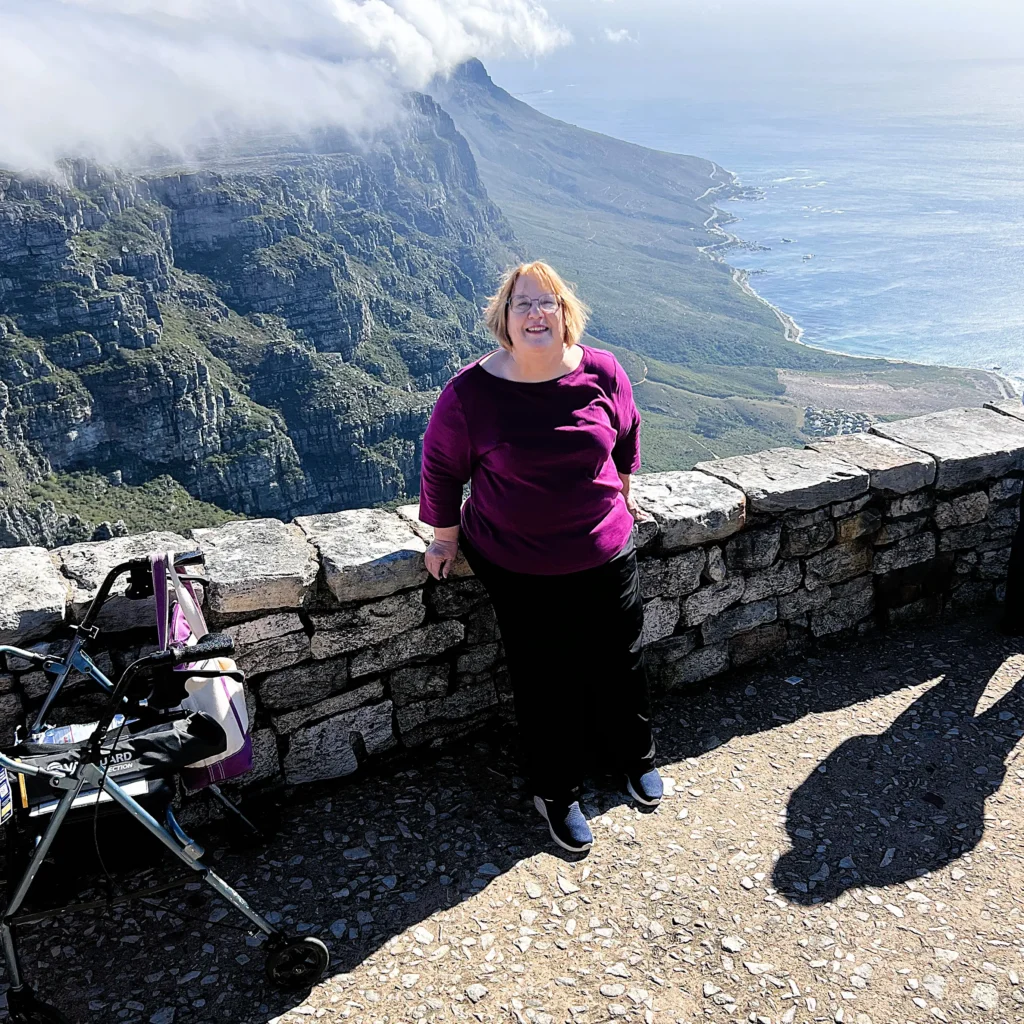 A middle-aged woman with light skin, short reddish-brown hair, and glasses is standing and smiling at the camera while leaning against a stone wall at a scenic viewpoint. She is wearing a purple long-sleeve top, black pants, and blue sneakers. To her left, a walker with a bag attached is resting against the wall. The background showcases a breathtaking mountainous landscape with cliffs covered in green vegetation, a coastline with a winding road, and the ocean stretching into the horizon. Low-hanging clouds partially cover the peaks, and the sunlight casts sharp shadows on the ground. The location appears to be a famous natural viewpoint, possibly Table Mountain in South Africa.