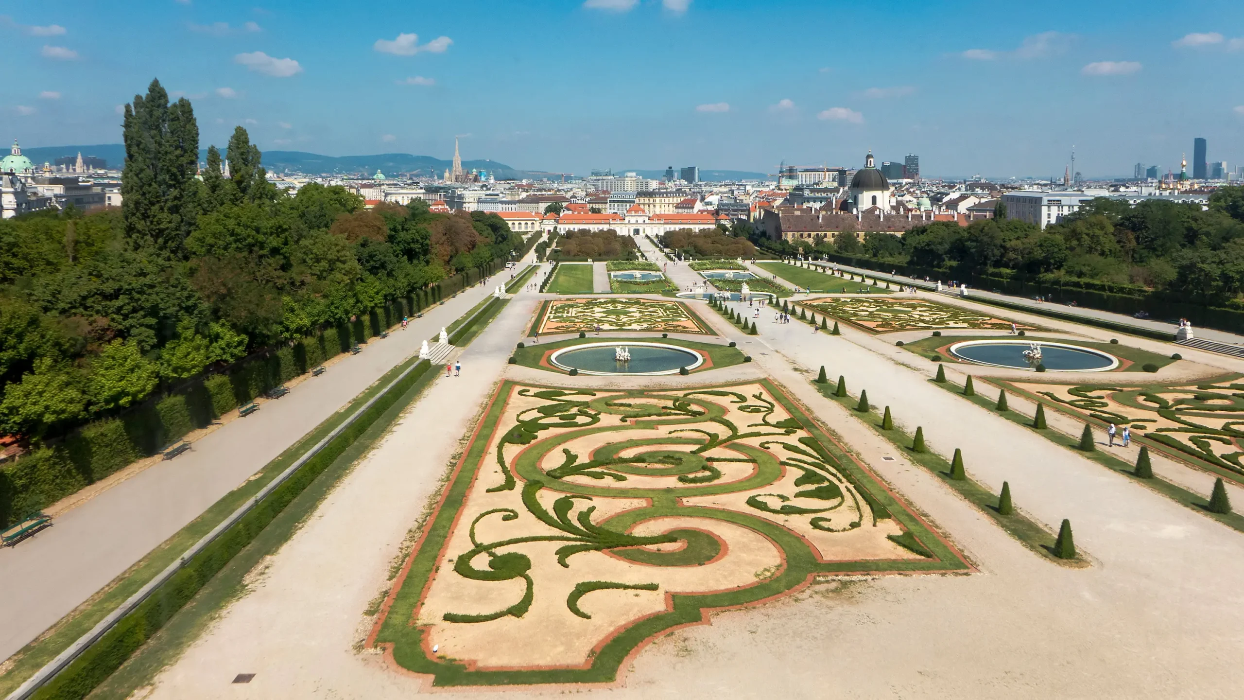 A scenic view of the gardens of Belvedere Palace in Vienna, Austria, featuring intricate symmetrical landscaping, decorative flower beds, and fountains. The city skyline with historic buildings and church spires is visible in the background under a bright blue sky. Visitors are strolling along the pathways, enjoying the well-maintained greenery.