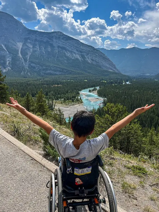A child in a wheelchair joyfully stretches their arms towards a stunning scenic view of a winding river, mountains, and lush forests.