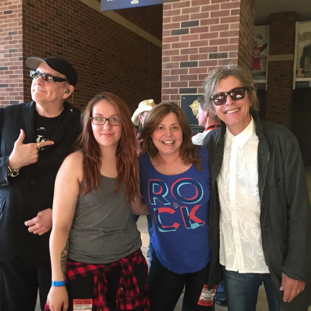 Four people pose together in a venue, wearing casual outfits and smiling, with memorabilia visible in the background.