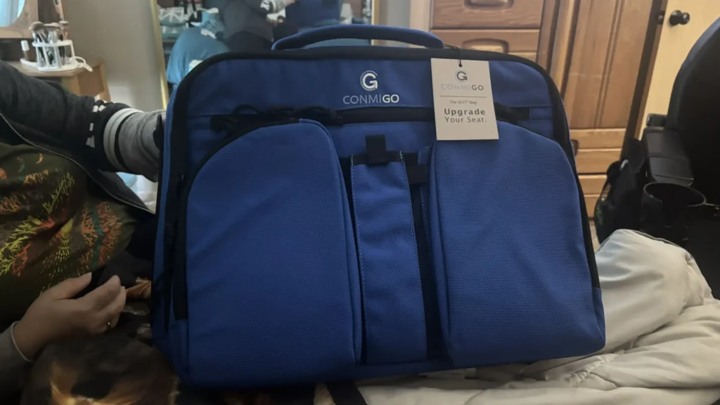 Blue travel bag labeled "CONMIGO The ULTR Bag" with a tag that reads "Upgrade Your Seat," resting on a bed.
