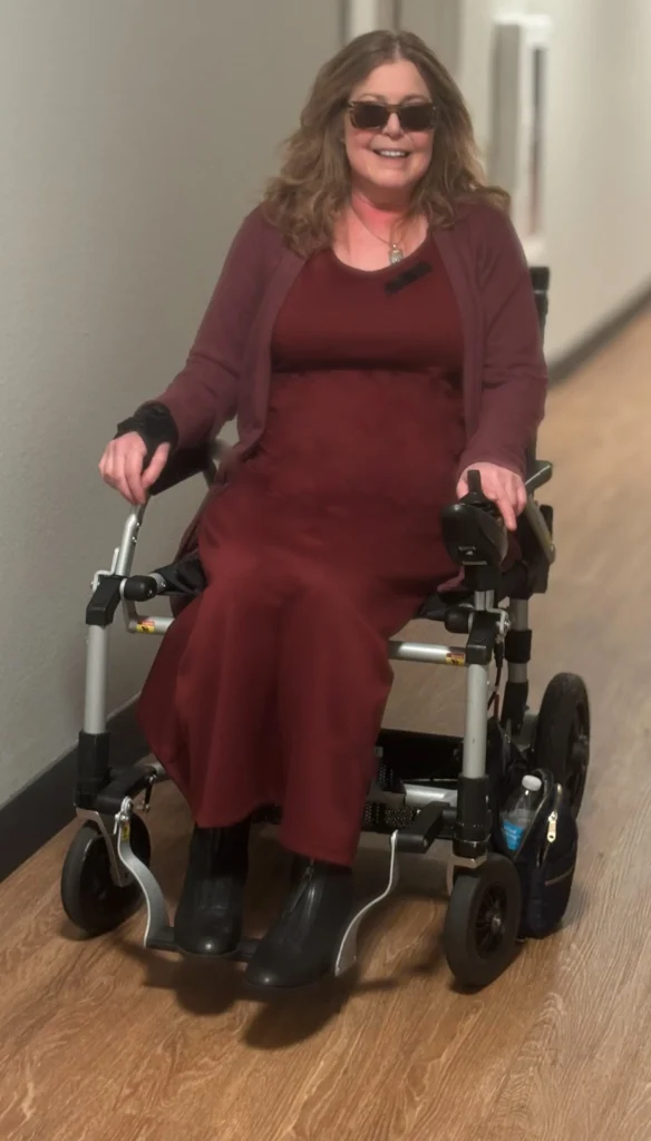 A woman in a flowing burgundy dress sits in a modern wheelchair on a wooden floor, with soft, natural lighting illuminating the hallway.