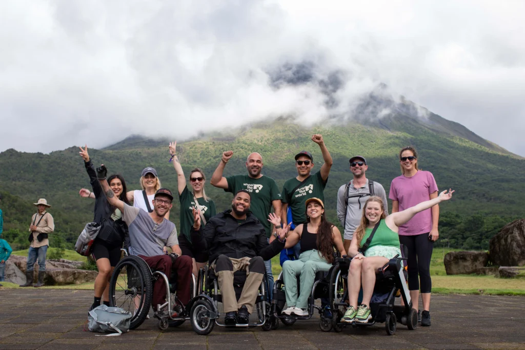 A diverse group of individuals in wheelchairs and standing, joyfully posing outdoors with a mountainous backdrop and cloudy skies.