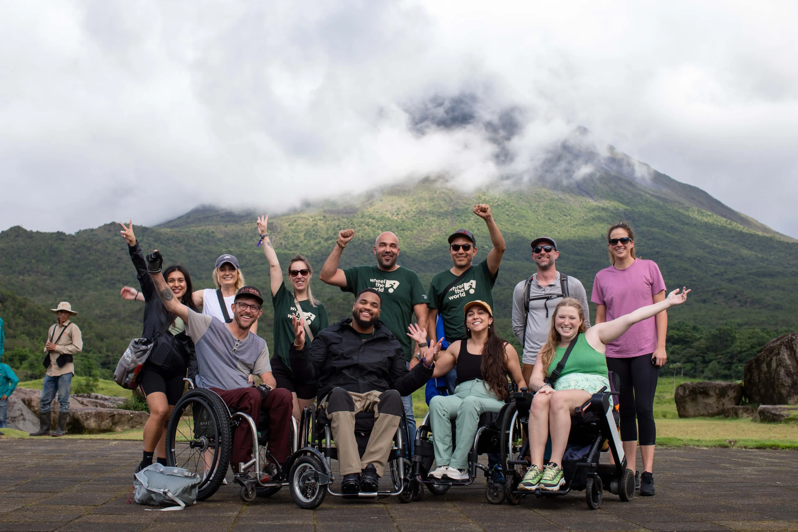 A diverse group of individuals in wheelchairs and standing, joyfully posing outdoors with a mountainous backdrop and cloudy skies.
