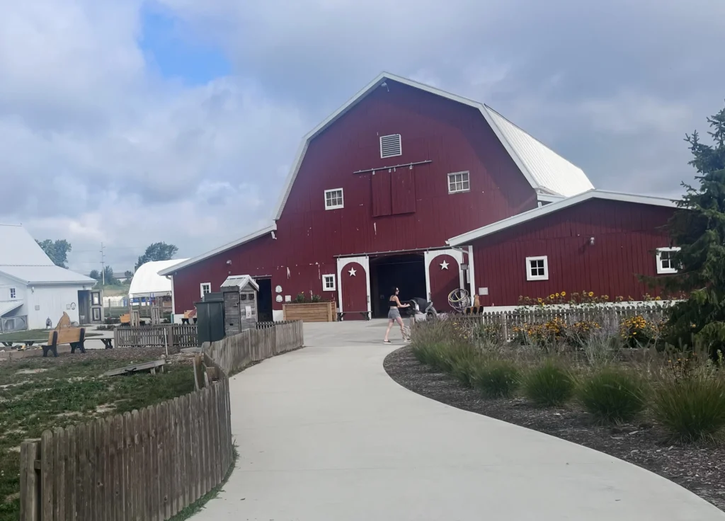 A woman walks along a paved path towards a large red barn surrounded by greenery and flowers under a cloudy sky.