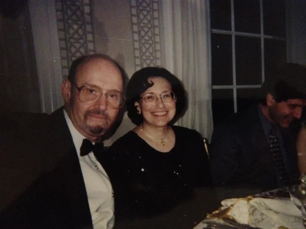 A formal dining scene with elegantly dressed individuals at a table, featuring white tablecloths and dim lighting.