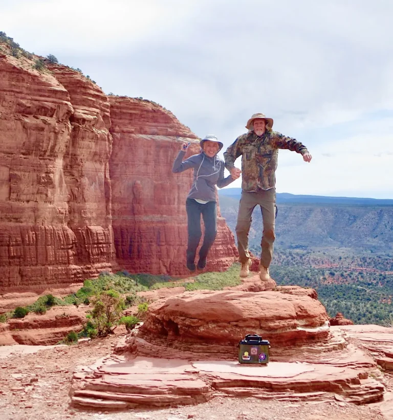 A couple joyfully jumps on a scenic hiking trail in Sedona, Arizona, surrounded by stunning red rock formations and expansive desert views. A green cooler with stickers is placed on the rocky surface, capturing the adventurous spirit of outdoor exploration.