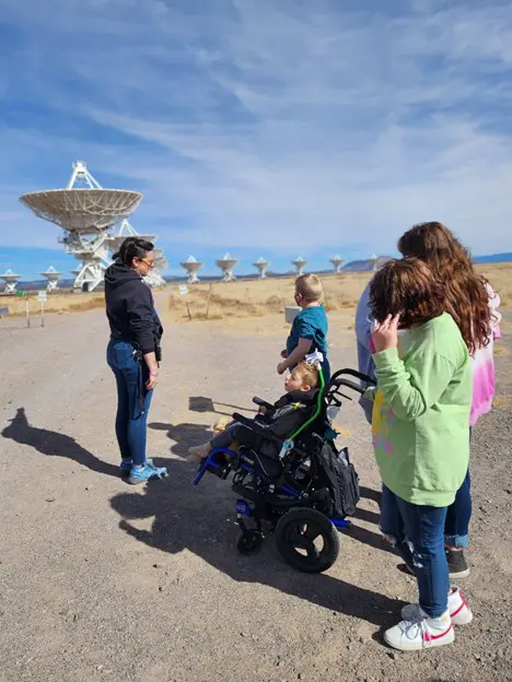 A group of four people, including a child in a stroller, stands near large satellite dishes under a blue sky.