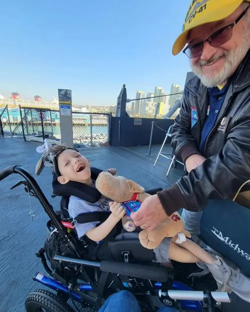 A child in a wheelchair holds a stuffed dog while an adult offers support, with a scenic view of a harbor in the background.