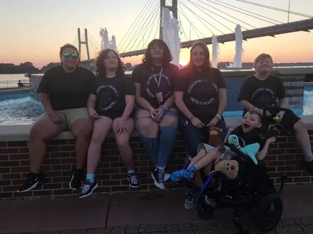 A group of five individuals sits on a brick ledge near a fountain at sunset, wearing matching black t-shirts, with a bridge in the background.