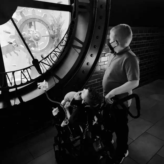 A boy in a mask watches fish through a large clock window, while a baby doll rests in a stroller beside him. Black and white image.