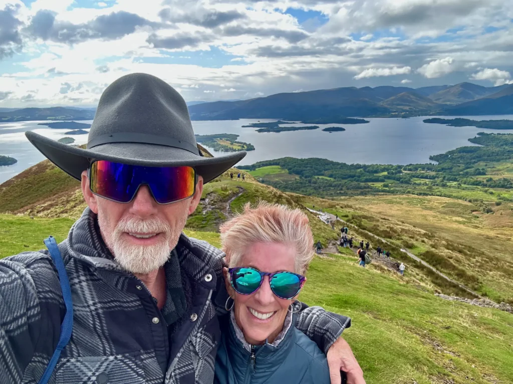 A man and woman smiling while taking a selfie on a mountain peak, with a scenic view of the landscape in the background.