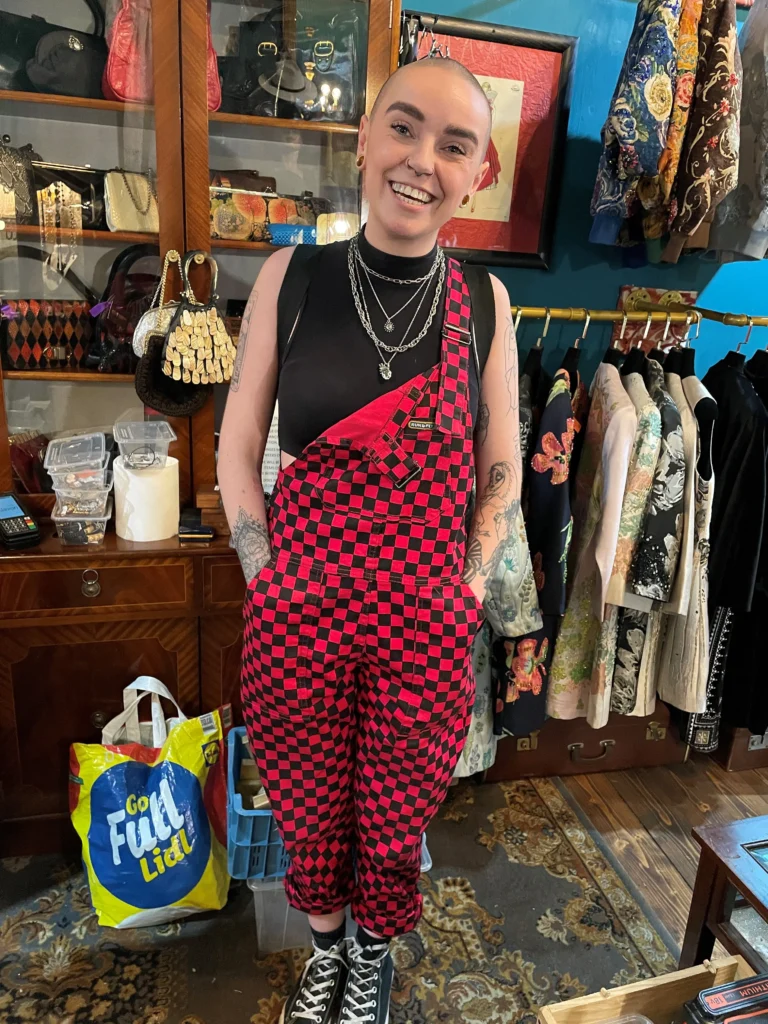 A person in red and black checkered overalls stands in a vintage clothing store, showcasing eclectic fashion items displayed behind them.