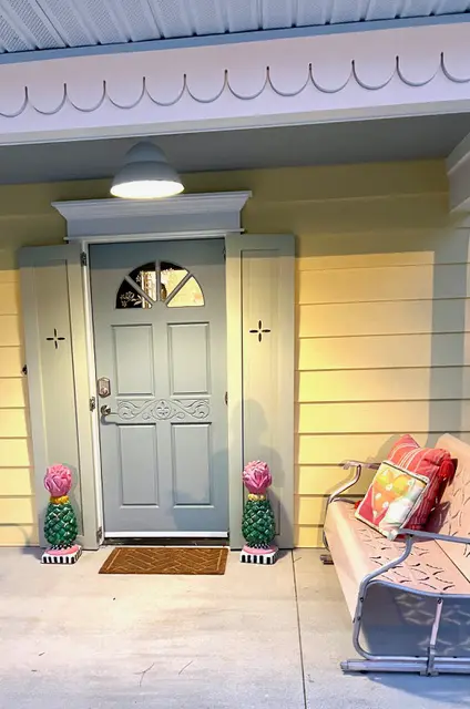 Charming entryway featuring a light blue door, colorful pineapple planters, and a pink bench with decorative pillows against a yellow wall.