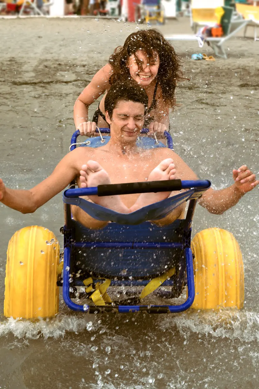 A young man sits in a blue water cart with yellow wheels, splashing through shallow water while being playfully pushed by a woman behind him.