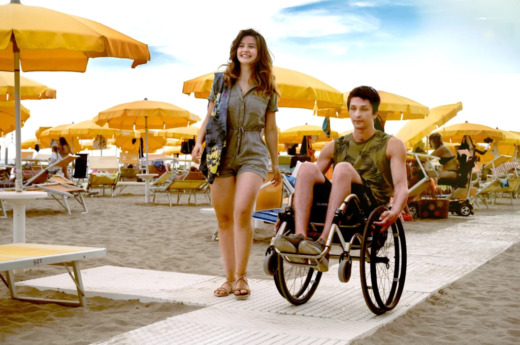 A woman in a romper walks alongside a man in a wheelchair at a sandy beach with yellow umbrellas and lounge chairs in the background.