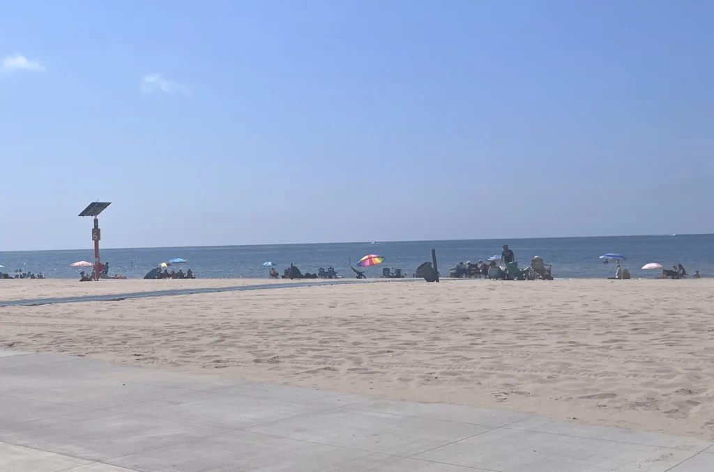 A sunny beach scene with people relaxing under colorful umbrellas, sandy shores, and calm blue ocean waters under a clear sky.