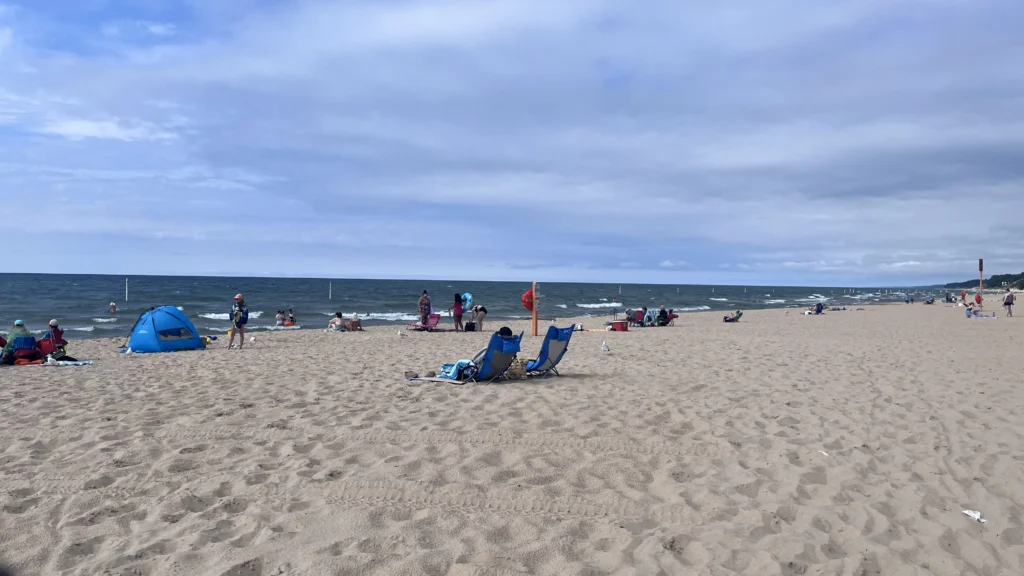 A sandy beach scene with people sunbathing, playing, and swimming in the waves under a cloudy sky, with colorful beach tents and chairs.