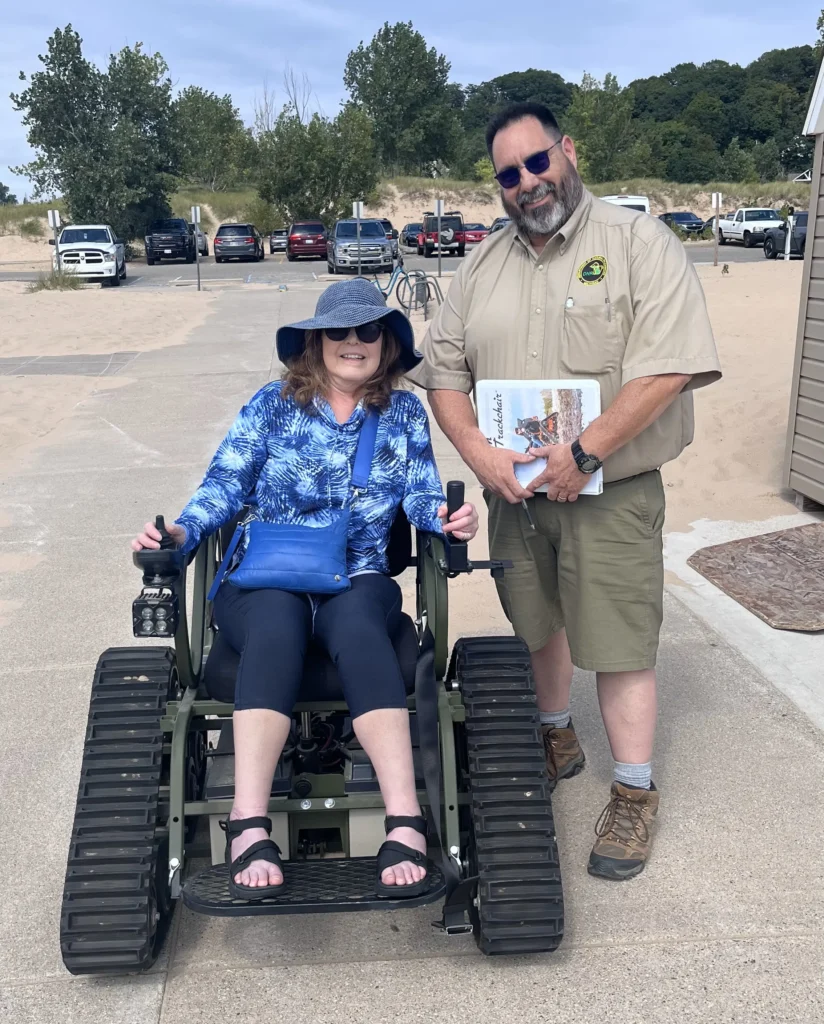 A woman sits in an all-terrain wheelchair, wearing a blue patterned jacket and a sunhat, alongside a man in a tan uniform holding a booklet.