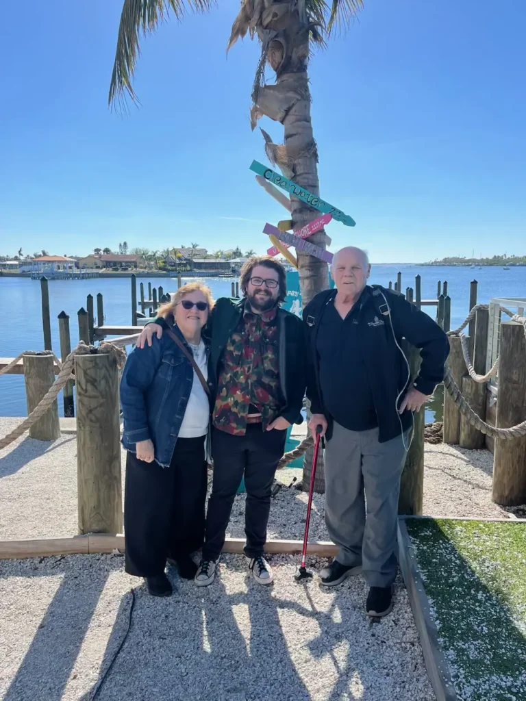 Three individuals pose for a photo in front of a palm tree with colorful directional signs by a waterfront on a sunny day.